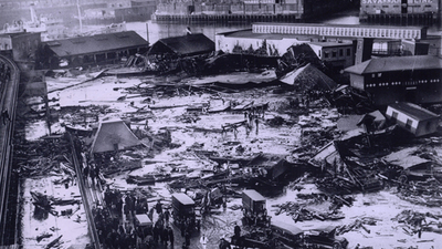 Twenty-one people were killed on Commercial Street in the North End when a tank of molasses ruptured and exploded. An 8-foot wave of the syrupy brown liquid moved down Commercial Street at a speed of 35mph. Wreckage of the collapsed tank visible in background, center, next to light-colored warehouse. Elevated railway structure visible at far left and the North End Park bathing beach to the far right.