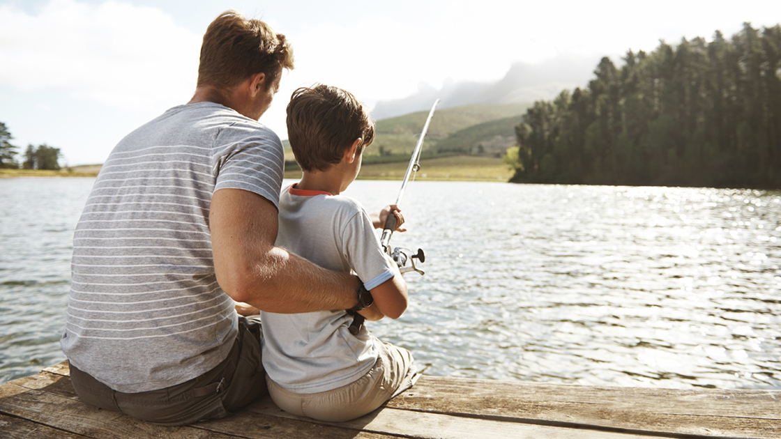 Rear view of a father trying to teach his son how to fish with a fishing rod by a lake