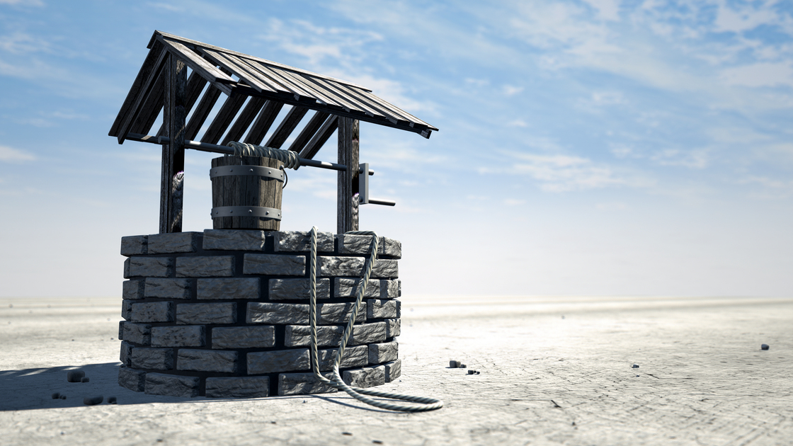 A brick water well with a wooden roof and bucket attached to a rope in a flat barren landscape with a blue sky background