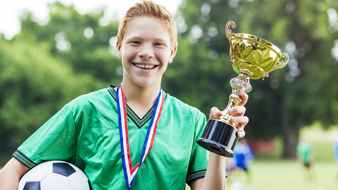 Proud male teenage Caucasian soccer player holds up the tournament winning trophy after game. He is also wearing a medal around his neck and holding a soccer ball. He has red hair and is wearing a green uniform. Soccer players are in the background.