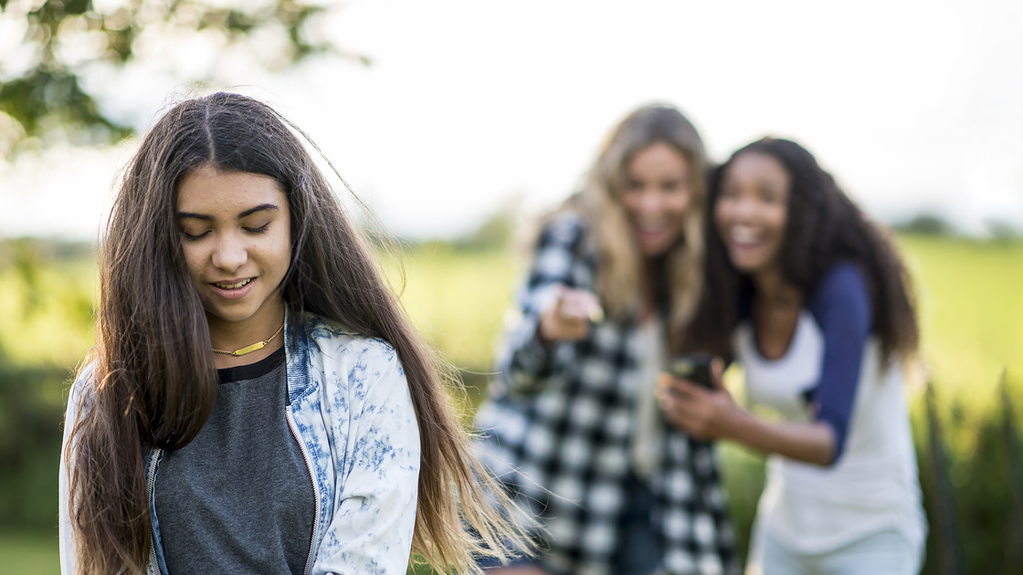 A multi-ethnic group of teenagers are outdoors on a sunny day. They are wearing casual clothing. Two girls are laughing at another girl in the foreground. She looks frustrated.