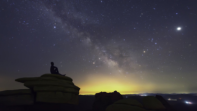 Man sitting on the rocks in solitude under the cover of dark, clear sky and looking at Milky Way galaxy while thinking about his existence.